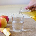 A woman’s hand pours apple cider vinegar in a glass bottle on a light background into a glass of water. Malic acid is beneficial for health and is used in cooking.